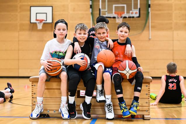 Vier Kinder sitzen auf einer Bank in der Turnhalle, halten Basketballs und lachen. Im Hintergrund sind weitere Kinder aktiv.