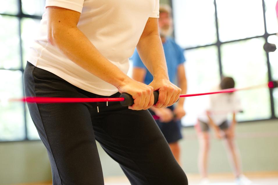 Nahaufnahme einer Person mit einem Widerstandsband im Fitnessstudio, umgeben von weiteren trainierenden Personen.