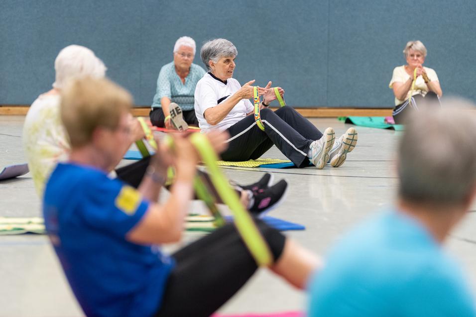 Senioren nehmen an einer Fitnessübung mit Fitnessbändern in einer Sporthalle teil, sitzend auf Fitnessmatten.