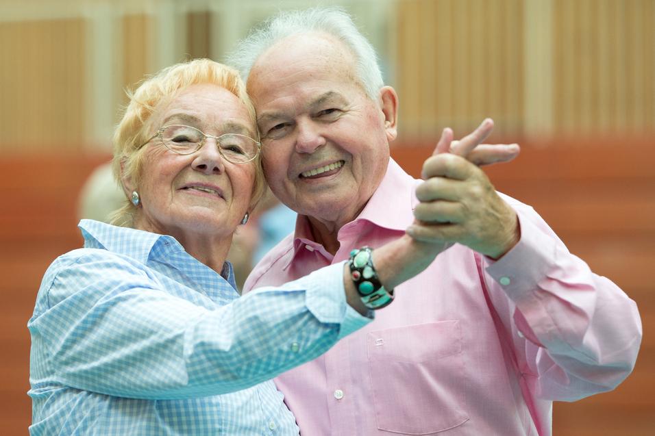 Ein älteres Paar umarmt sich lächelnd und tanzt in einer Sporthalle. Ihre Kleidung ist hell und farbenfroh.