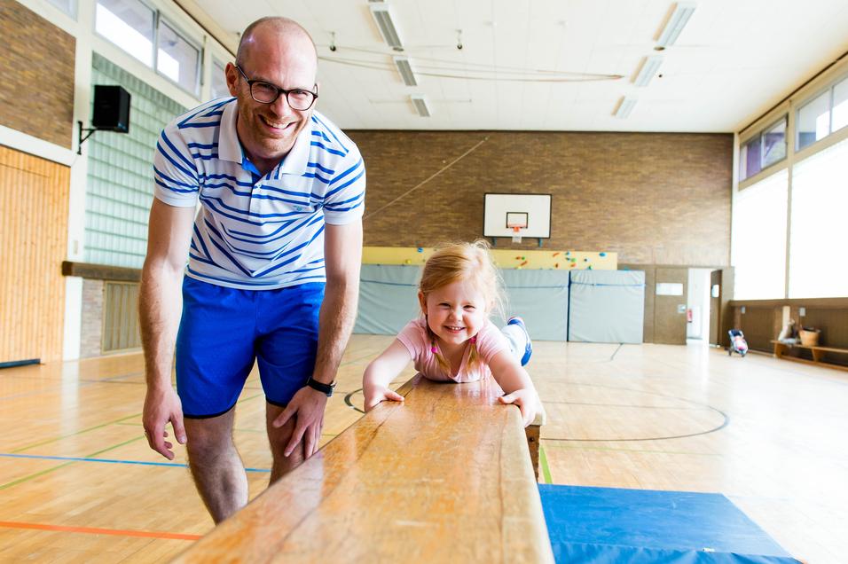 Mann lehnt am Schwebebalken, während ein lächelndes Mädchen darauf liegt, in einer hellen Sporthalle.