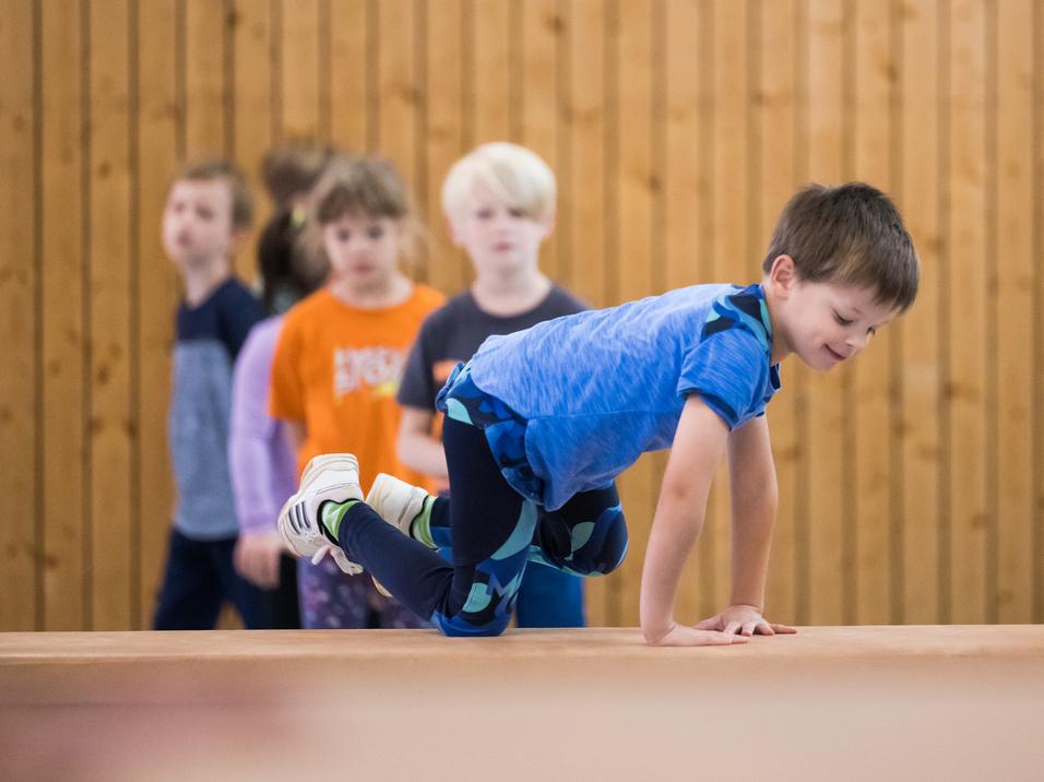 Ein Junge balanciert auf einem Blickkasten, während mehrere Kinder im Hintergrund zuschauen. Holzpaneele sind sichtbar.