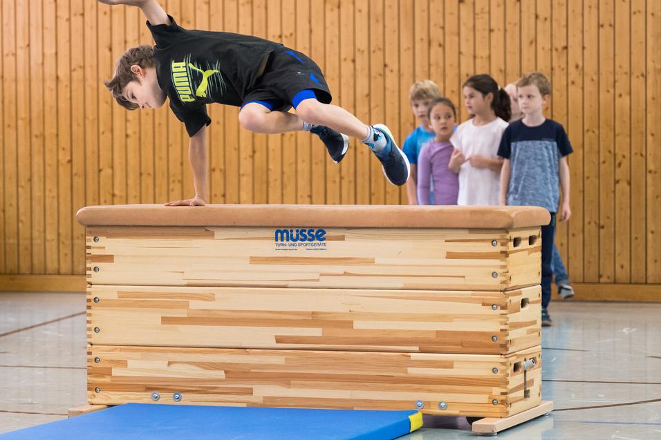 Ein Junge springt über ein Turngerät, während eine Gruppe Kinder und ein Trainer zuschauen. Sporthalle mit Holzverkleidung.