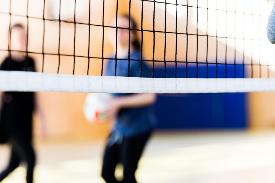 Unscharfe Silhouetten von zwei Personen hinter einem Volleyballnetz in einer Sporthalle. Eine Person hält einen Volleyball.
